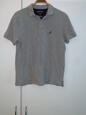 Grå pikétröja från Nautica - Klassisk grå pikétröja från Nautica i 100% bomull. Tröjan har korta ärmar, krage och knappar framtill samt en liten broderad logga på bröstet. Perfekt till en avslappnad och stilren look.