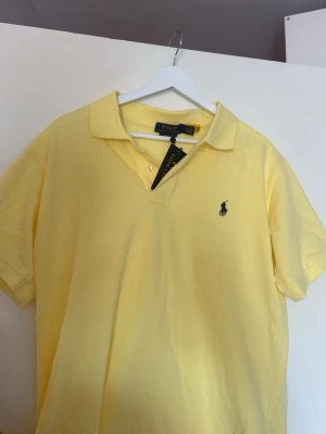 Gul pikétröja från Polo Ralph Lauren - Klassisk gul pikétröja från Polo Ralph Lauren med korta ärmar och den ikoniska broderade loggan på bröstet. Tröjan har krage och knappar vid halsen. Perfekt för en avslappnad och stilren look.