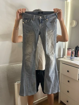 Bootcut jeans från True Religion - Snygga ljusblå bootcut jeans från True Religion med klassiska detaljer och kontrastsömmar. Jeansen har fem fickor, silverfärgade knappar och en ikonisk bakficka med lock och broderad logga. Perfekta för en avslappnad och trendig look.
