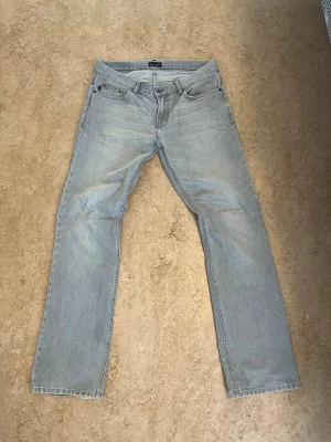 Gråa bootcut jeans - Säljer ett par ljusgrå jeans med bootcut passform och klassisk femficksdesign. Jeansen har normal midja och är tillverkade i denim med subtila slitningar för en avslappnad look. Perfekta till vardags för en enkel och stilren outfit.