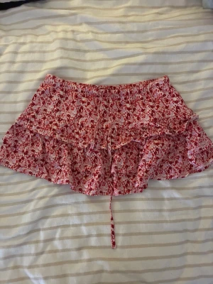 Rosa blommig volangkjol med snörning - Supersöt kort kjol i rosa med rött blommönster och volanger. Kjolen har en smockad midja och snörning framtill, vilket ger en avslappnad och trendig look. Perfekt för varma dagar och enkel att matcha med en enkel topp.