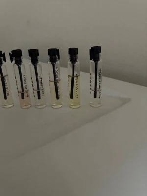 Set med parfymprover i glasflaskor - Säljer ett set med sex små parfymprover i genomskinliga glasflaskor med svarta spraykorkar. Varje flaska har en etikett med parfymens namn och innehåller olika nyanser av ljusgul och klar vätska. Perfekt för dig som vill testa olika dofter.