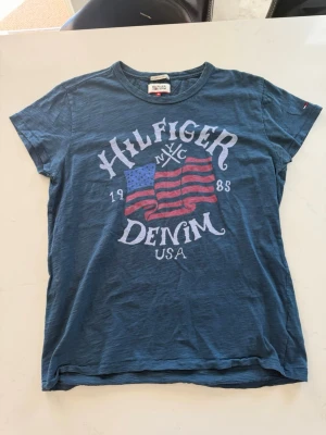 Blå Hilfiger Denim t-shirt - Snygg blå t-shirt från Hilfiger Denim med stort tryck framtill som visar amerikansk flagga och texten 'Hilfiger Denim NYC 1985 USA'. Klassisk rund hals och korta ärmar. Perfekt för en avslappnad och cool stil.