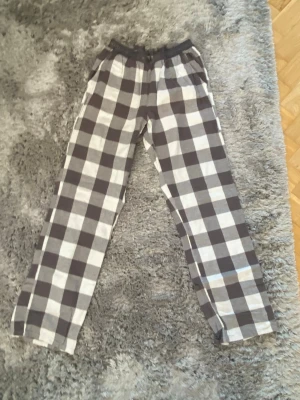 Rutiga pyjamasbyxor i bomull - Säljer ett par klassiska pyjamasbyxor med svart och vitt rutmönster. Byxorna har resår i midjan och raka ben, perfekta för chill hemma. Materialet känns mjukt och skönt, troligen bomull. Passar dig som gillar enkel och avslappnad stil.