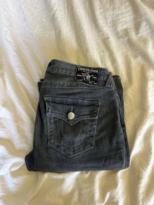 Grå True Religion jeans - Snygga grå jeans från True Religion med klassiska kontrastsömmar och detaljerade bakfickor med lock och silverfärgade knappar. Jeansen har en rak passform och är tillverkade i mjukt denimtyg. Perfekta för en avslappnad och trendig look.