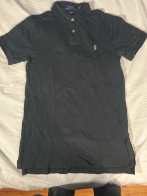 Svart pikétröja från Polo Ralph Lauren - Klassisk svart pikétröja från Polo Ralph Lauren med grå broderad logga på bröstet. Tröjan har korta ärmar, krage och två knappar framtill. Tillverkad i mjuk bomull och har en slim fit passform som sitter snyggt.