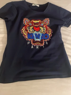 Mörkblå Kenzo t-shirt med tiger - Ifrån Dubai Snygg mörkblå t-shirt från Kenzo med ikoniskt broderat tigermotiv i flera färger och texten 'KENZO PARIS' framtill. T-shirten har rund hals och korta ärmar. Perfekt för dig som vill sticka ut med en färgstark och trendig look.
