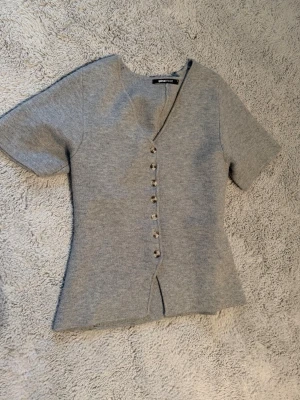 Grå v-ringad topp från Gina Tricot - Snygg grå topp från Gina Tricot med v-ringning och korta ärmar. Toppen har dekorativa knappar framtill och är lätt figursydd för en smickrande passform. Perfekt att styla med jeans eller kjol för en trendig look.
