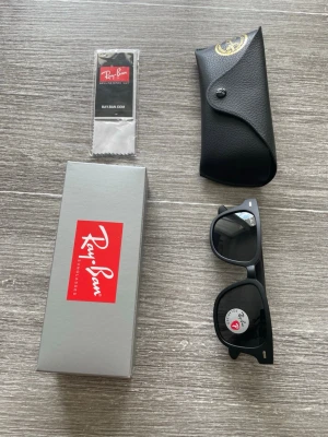 Svarta Ray-Ban Wayfarer solglasögon - Klassiska svarta Ray-Ban Wayfarer solglasögon med mörka glas och ikonisk fyrkantig form. Kommer med originalfodral, putsduk och box. Perfekta för dig som vill ha en tidlös och cool look i solen.