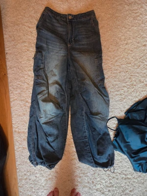 Baggy cargobyxor i denim - Säljer ett par baggy cargobyxor i blå denim med stora sidofickor och dragsko vid benslut. Byxorna har resår i midjan bak och en bakficka. Perfekta för en avslappnad streetstil.