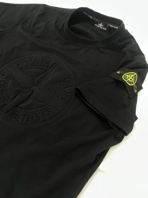  Stone Island  - Snygg svart t-shirt från Stone Island med stor ton-i-ton broderad logga på bröstet och den klassiska patchen på ärmen. T-shirten har rund hals och är tillverkad i mjuk bomull. Perfekt för dig som gillar streetwear och vill ha något stilrent men ändå med edge.