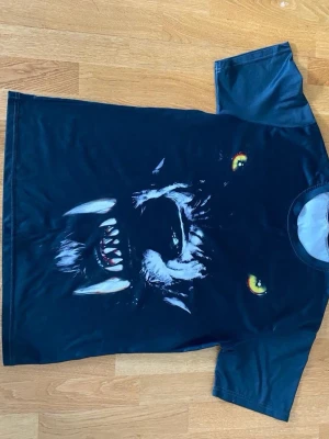 Svart t-shirt med vargtryck - Cool svart t-shirt med ett stort tryck av en varg med lysande gula ögon och öppet gap på framsidan. T-shirten har korta ärmar och rund hals. Perfekt för dig som gillar djurmotiv och vill sticka ut med en unik design.