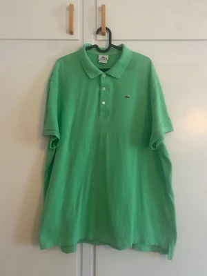 Grön pikétröja från Lacoste - Snygg grön pikétröja från Lacoste med klassisk krage och knappar framtill. Tröjan har korta ärmar och den ikoniska krokodilloggan på bröstet. Perfekt för en avslappnad och stilren look.