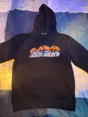 Svart hoodie med tryck och huva - Snygg svart hoodie med stor huva och dragsko. Framtill finns ett färgglatt tryck med texten 'Shooters' och sportmotiv i orange och vitt. Hoodien har en klassisk magficka och ribbade muddar vid ärmslut och nederkant. Perfekt för en avslappnad och cool stil.