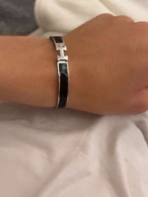 Svart armband  - Snyggt svart armband  med silverfärgade detaljer och klassiskt H-spänne. Armbandet har en stilren och elegant design i metall och emalj, perfekt för dig som gillar exklusiva accessoarer. Kommer i original orange box