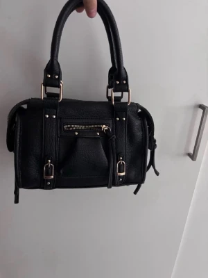 Svart handväska med detaljer - Snygg svart handväska i skinnimitation med guldfärgade metalldetaljer, dubbla handtag och en liten ficka med dragkedja framtill. Väskan har spännen och dekorativa remmar som ger en cool look.
