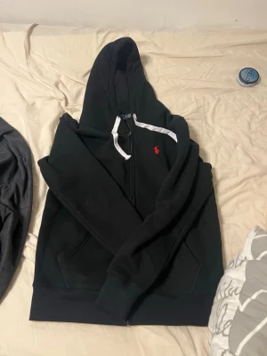 Svart hoodie med dragkedja och huva - Säljer en svart hoodie från Polo Ralph Lauren med dragkedja framtill, vit dragsko i huvan och röd logga broderad på bröstet. Hoodien har kängurufickor och ribbade muddar vid ärmslut och nederkant. Perfekt för en chill och stilren look.