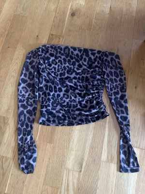 Leopardmönstrad mesh topp offshoulder - Säljer en trendig offshoulder topp i mesh med leopardmönster i svart och grått. Toppen har lång ärm och är croppad med snygga rynkade detaljer på sidan. Perfekt för dig som vill sticka ut med en edgy look.