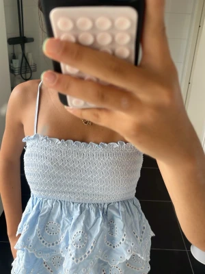 Ljusblå bandeau topp från Stradivarius - Supersöt ljusblå bandeau topp från Stradivarius med smockad överdel och tunna axelband. Toppen har broderade detaljer och volanger nedtill som ger en romantisk vibe. Perfekt för varma dagar och sommarens alla äventyr.
