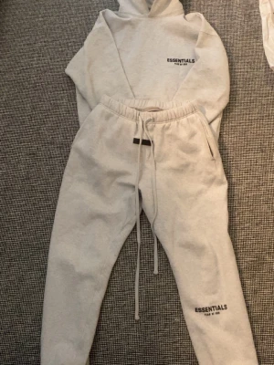 Grå Essentials hoodie och byxor - Säljer ett set med grå hoodie och matchande sweatpants från Essentials Fear of God. Hoodien har tryck på bröstet och byxorna har samma tryck på benet. Båda plaggen är i mjukt och bekvämt material, perfekta för chill dagar hemma.