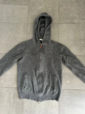 Mörkgrå hoodie med dragkedja - Mysig mörkgrå hoodie med dragkedja framtill och två fickor. Tillverkad i ett mjukt, stickat material som känns skönt mot huden. Huva med dragsko och ribbade muddar vid ärmslut och nederkant. Perfekt för en avslappnad och chill stil. Tröjan har ett pyttelitet hål som är på armbågen med det går mycket lätt att sy ihop! Bara att höra av sig vid funderingar