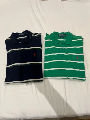 Randiga pikétröjor från Ralph Lauren - Två klassiska pikétröjor från Ralph Lauren i storlek M. En är marinblå med vita och gröna ränder, den andra är grön med vita ränder. Båda har krage, knappar och den ikoniska broderade loggan på bröstet. Perfekta för en avslappnad men stilren look. Pris 150kr styck vill man köpa båda så kostar de 300kr hör bara av er vad ni vill köpa.