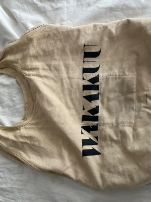 Beige shoppingväska med tryck - Snygg beige shoppingväska i kraftig bomull med svart texttryck på framsidan. Väskan har rundade handtag och en enkel, stilren design som passar perfekt till vardags. Praktisk och rymlig för alla dina grejer.