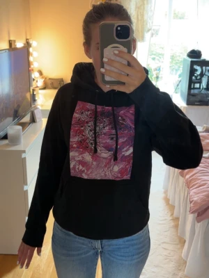 Svart hoodie The Cool Elephant - Säljer en svart hoodie från The Cool Elephant med en stor färgstark grafisk print i rosa och rött framtill. Hoodien har huva med svarta snören och en liten logga tryckt på ryggen. 