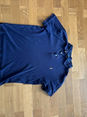 Marinblå piké från Polo Ralph Lauren - Snygg marinblå pikétröja från Polo Ralph Lauren  i ett stretchigt matriall som gör den perfekt både till vardags eller på golfbanan. Den har ett minimalt hål som man ser på bild nr 3 men annars är allt prima!