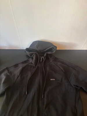 Svart hoodie med dragkedja från BOSS - Snygg svart hoodie från BOSS med dragkedja framtill och justerbar huva med snören. Diskret BOSS-logga på bröstet och mjukt material som känns skönt mot huden. Perfekt för en avslappnad och stilren look.
