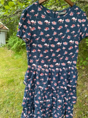 Mörkblå klänning med flamingomönster - Supersöt mörkblå klänning från Oasis med korta ärmar och ett lekfullt mönster av rosa flamingos och blommor. Klänningen har rund halsringning, markerad midja och faller i flera lager för en avslappnad look. Perfekt för sommarens alla tillfällen! Märkt ned 14, men passar mig som har 38/M