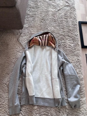 Grå hoodie från Burberry med mönstrad huva - Säljer en grå hoodie från Burberry med dragkedja och klassiskt rutmönster i beige, brunt och rött i huvan. Insidan är mjuk och fleece-liknande, perfekt för chill dagar. Hoodien har dragsko i huvan och breda muddar vid ärmar och nederkant.
