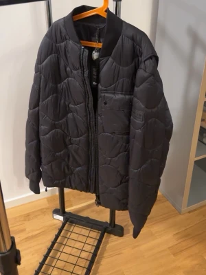 Svart pufferjacka från Barbour - Snygg svart pufferjacka från Barbour International med quiltat mönster och ribbad krage. Jackan har dragkedja framtill, en bröstficka och Barbour-logga på ärmen. Perfekt för kalla dagar och ger en modern look. Använts 0 gånger.