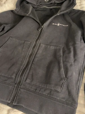 Svart hoodie från Sail Racing - Svart hoodie med dragkedja från Sail Racing. Klassisk modell med två stora fickor framtill, huva med dragsko och diskret logga på bröstet. Mjuk insida och detaljerade sömmar vid huvan. Perfekt för en avslappnad och sportig stil.