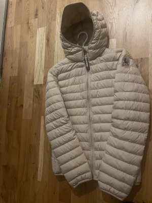 Ljusbeige dunjacka med huva - Snygg ljusbeige dunjacka med quiltad design och fast huva. Jackan har dragkedja framtill och en logotyp på vänster ärm. Perfekt för kalla vinterdagar och ger en stilren look. Lätt och smidig att bära, passar till det mesta.