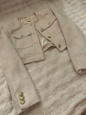 Beige bouclé cardiganjacka från Amisu - Stilren beige bouclé cardiganjacka från Amisu med rund halsringning, dekorativa guldknappar och två stora bröstfickor. Jackan är fodrad med glansigt tyg och har en croppad passform som ger en trendig look.