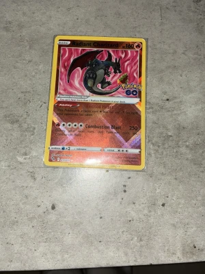 Radiant Charizard Pokémonkort - Radiant Charizard Pokémonkort från Pokémon GO-serien. Kortet har en röd och gul ram med holografisk effekt och visar Charizard med svarta vingar mot en eldig bakgrund. Perfekt för samlare eller Pokémonfans.