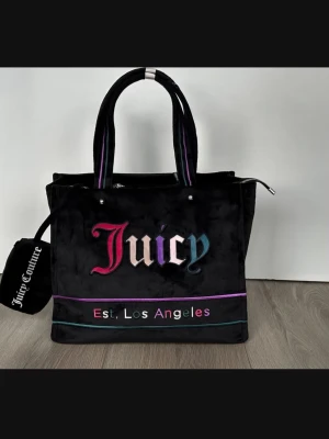 Juicy Couture svart shoppingväska - En mycket rymlig Tote Bag shoppingväska i svart sammet från Juicy Couture med med tillhörande plånbok. Färgglada broderade detaljer och texten 'Juicy' samt 'Est. Los Angeles' på framsidan. Väskan har dubbla handtag, dragkedja upptill och dekorativa ränder i lila och turkos. Perfekt för att bära allt du behöver. Helt ny och oanvänd såklart.   Mått i cm: 38/30/15, se bild!