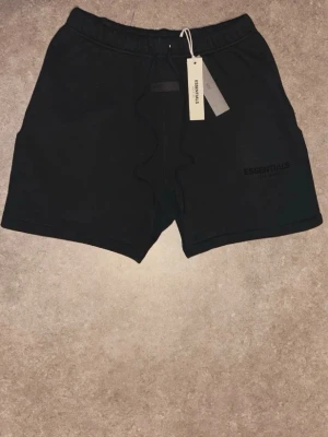 Svarta shorts från Essentials - Säljer ett par svarta shorts från Essentials Fear of God med snörning i midjan och diskret logga broderad på vänster ben. Shortsen har en avslappnad passform och är perfekta för sommaren. Materialet känns mjukt och bekvämt.