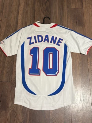 Frankrike Zidane 10 matchtröja - Säljer en vit Frankrike matchtröja från Adidas med Zidane och nummer 10 på ryggen. Tröjan har blå och röda detaljer, broderad landslagssköld och Adidas-logga på bröstet. Perfekt för fotbollsfans som vill sticka ut på planen eller läktaren.