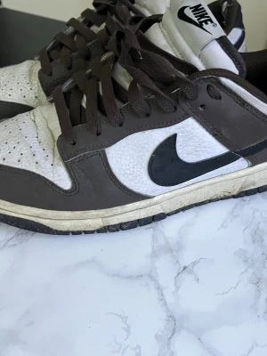 Nike Dunk Low brun och vit - Säljer ett par Nike Dunk Low sneakers i brunt och vitt med svart swoosh. Skorna har lågt skaft, bruna skosnören och vit mellansula. Ovandelen är i läder med perforerade detaljer framtill och klassisk Nike-logga på hälen.