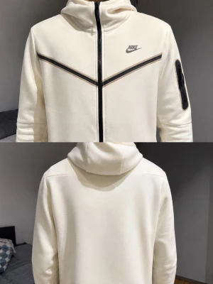 Vit Nike hoodie med dragkedja - Snygg vit hoodie från Nike med hel dragkedja framtill och svart Nike-logga på bröstet. Har en huva och svarta detaljer i form av linjer över bröstet samt en ficka med dragkedja på ärmen. Perfekt för en sportig och avslappnad stil. Men två lite fläckar 