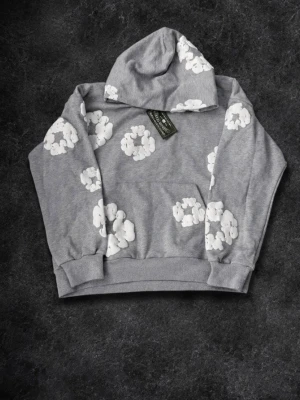 Grå hoodie med vita molntryck - Säljer en grå hoodie med huva och stora vita molnliknande tryck över hela plagget. Hoodien har en klassisk känguruficka framtill och ribbade muddar vid ärmslut och nederkant. Perfekt för dig som gillar en lekfull och avslappnad stil.