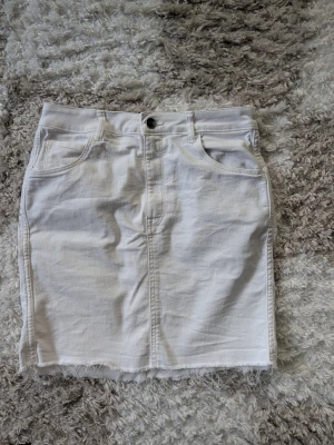 Vit jeanskjol med rå kant - Säljer en vit jeanskjol i kort modell med rå, fransig kant nedtill. Kjolen har klassiska fem fickor, bälteshällor och knappstängning framtill. Perfekt till sommaren för en avslappnad och trendig look.