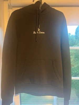 Svart hoodie från Ralph Lauren - Snygg svart hoodie från Ralph Lauren med vit broderad logga och text på bröstet. Klassisk känguruficka framtill, justerbar dragsko i huvan och ribbade muddar vid ärmar och nederkant. Perfekt för en avslappnad och stilren look. Kan skicka flera bilder
