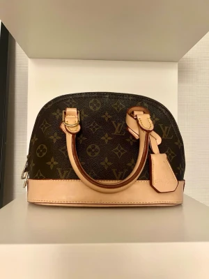 Vintage Alma Monogram väska - Snygg Alma handväska i klassiskt brunt monogramcanvas med beige läderdetaljer och dubbla handtag. Väskan har silverfärgade metalldetaljer och dragkedja upptill. Perfekt storlek för det viktigaste och ikonisk design.