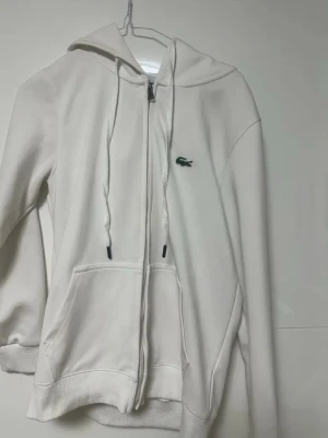 Vit zip-hoodie från Lacoste - Snygg vit hoodie med dragkedja från Lacoste. Klassisk krokodil-logga på bröstet, huva med dragsko och stora fickor framtill. Tillverkad i mjukt material som känns skönt mot huden. Perfekt för en clean och stilren look.