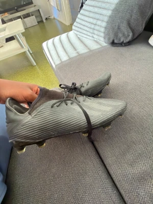 Grå fotbollsskor med dobbar - Säljer ett par svarta fotbollsskor från Adidas med snörning och ribbad ovansida. Skorna har fasta dobbar och en strumpliknande krage för extra stöd runt ankeln. Perfekta för spel på gräsplaner och ger bra grepp och kontroll på bollen.