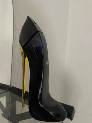 Carolina Herera  - Snygg parfymflaska formad som en högklackad sko i svart och guld. Flaskan har en elegant och modern design med glänsande yta och gulddetaljer. Perfekt som dekoration på sminkbordet eller i badrummet.