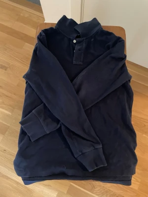 Mörkblå långärmad pike Polo Ralph Lauren - Klassisk mörkblå långärmad tröja från Polo Ralph Lauren med krage och två knappar vid halsen. Tröjan har en liten röd logga broderad på bröstet och ribbade muddar vid ärmslut och nederkant. Perfekt till en avslappnad men stilren look. Skulle behövas en tvätt i tvättmaskinen för lite smutsig om det är smuts som går bort. Står att den är large men skulle säga S